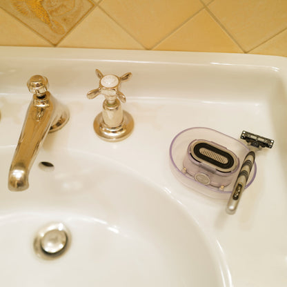 A clear Razor Rinser, which shoots water jets to unclog your razor, situated on a bathroom countertop next to a sink.